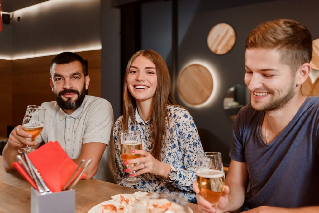 Two men and woman holding glasses of beer and sitting in pizzeria